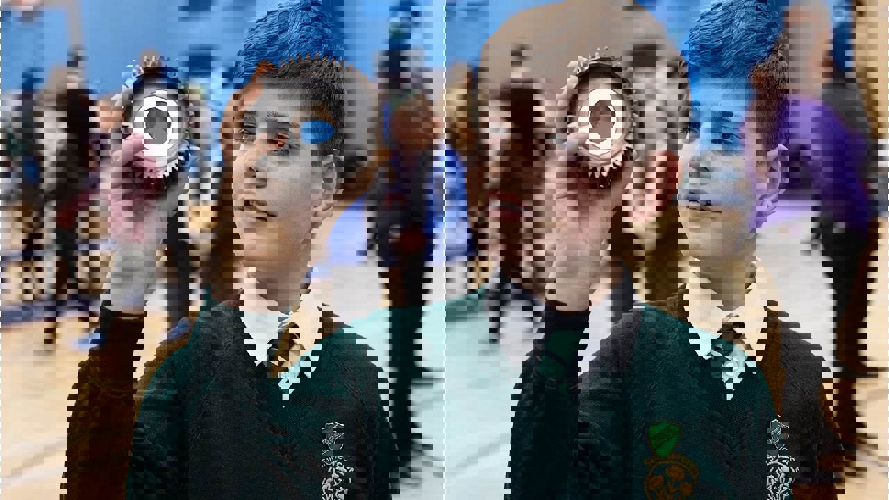 Young person in a school hall holding a piece of scientific equipment as other young people in the background work 