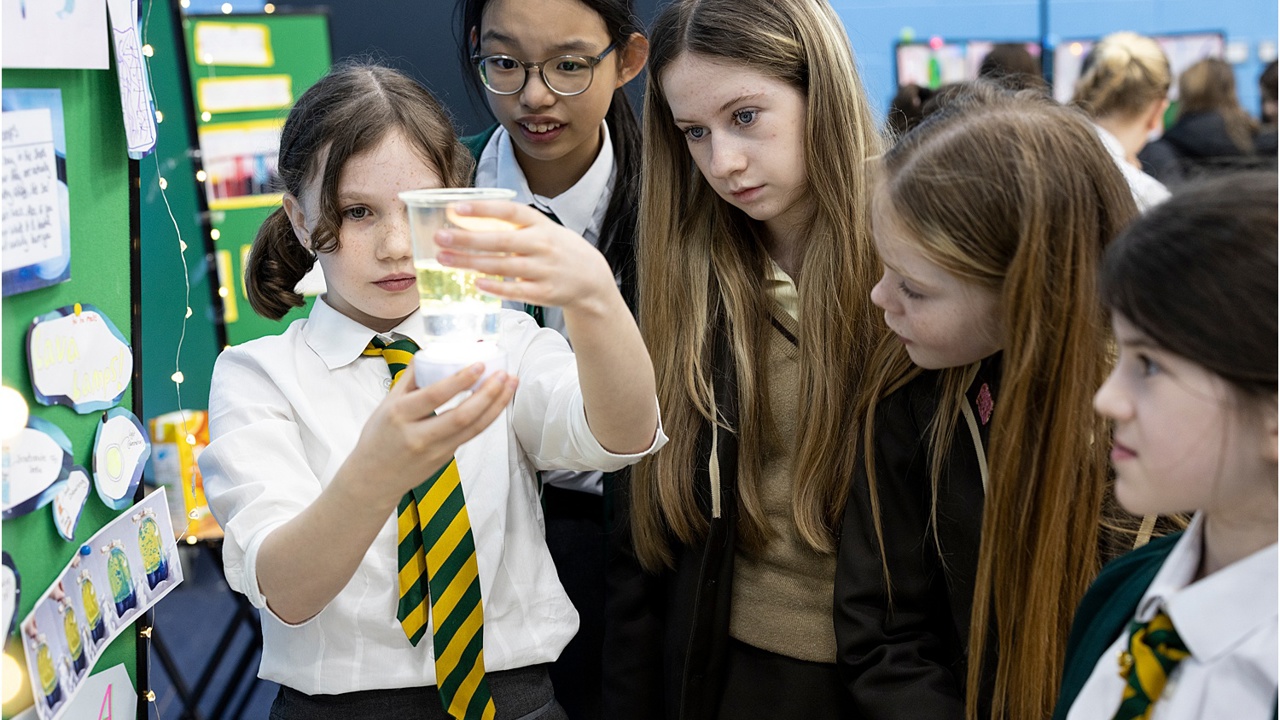 Five young people at a science fair, investigating a beaker of liquid. One holds the beaker as the others look on with interest.