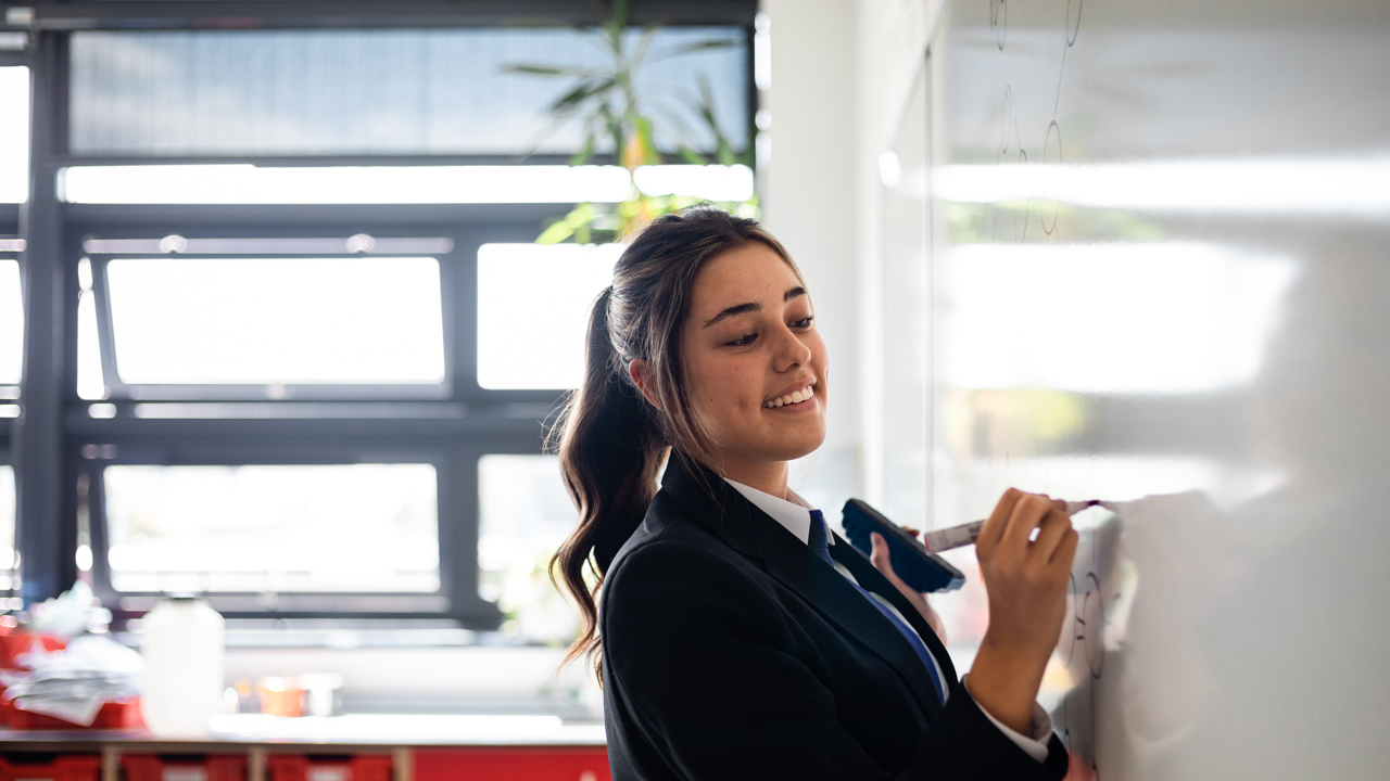 Secondary school student in a classroom writing on a whiteboard.