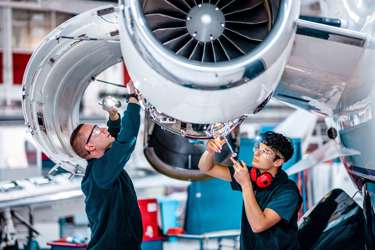 Two engineers working on an aircraft. They have protective wear and tools and are underneath a propeller. 