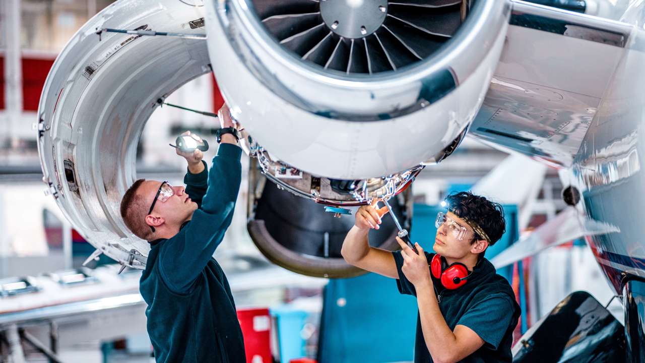 Two engineers working on an aircraft. They have protective wear and tools and are underneath a propeller. 