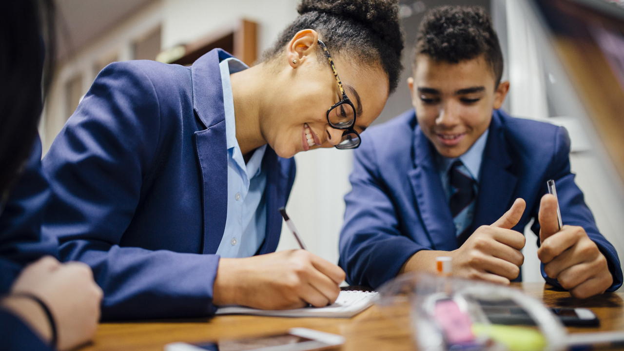 Secondary school students in a classroom at a desk working. One writes as the others look on.