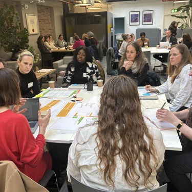 A working session in a meeting room, groups of people sit around tables discussing a topic
