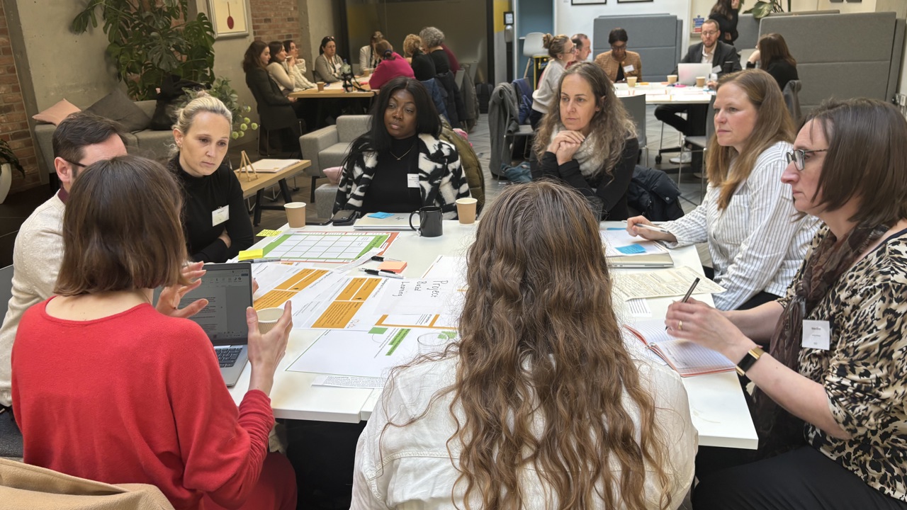 A working session in a meeting room, groups of people sit around tables discussing a topic