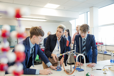 A school science lab where students are conducting an experiment with apparatus and protective equipment while a teacher looks on