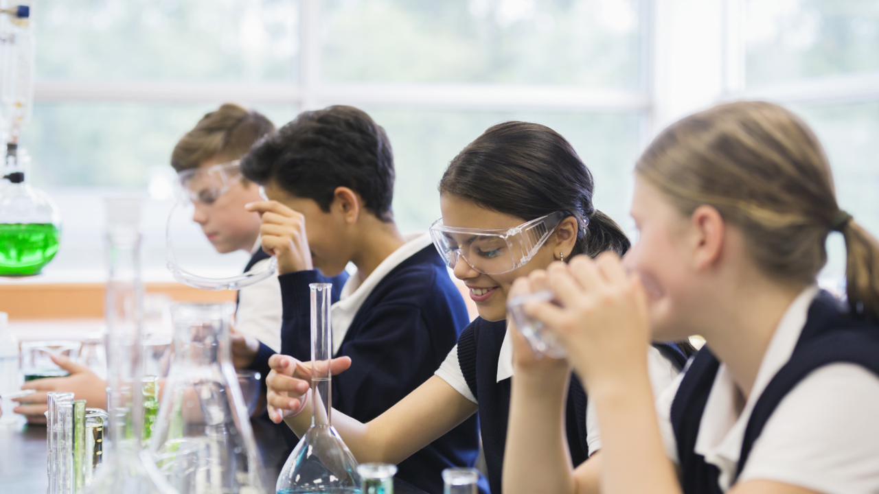 Four secondary school pupils working in a science lesson. They are all wearing protective goggles as they work with scientific equipment such as beakers, flasks and coloured liquids.