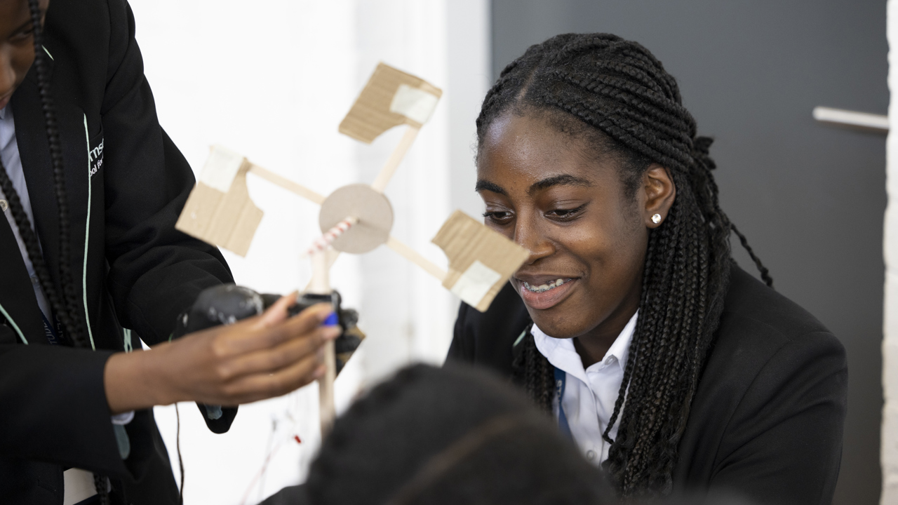 Close up shot of a group of young people building a model of a wind turbine in their science lesson. One young person is holding the model as the camera focuses on another smiling